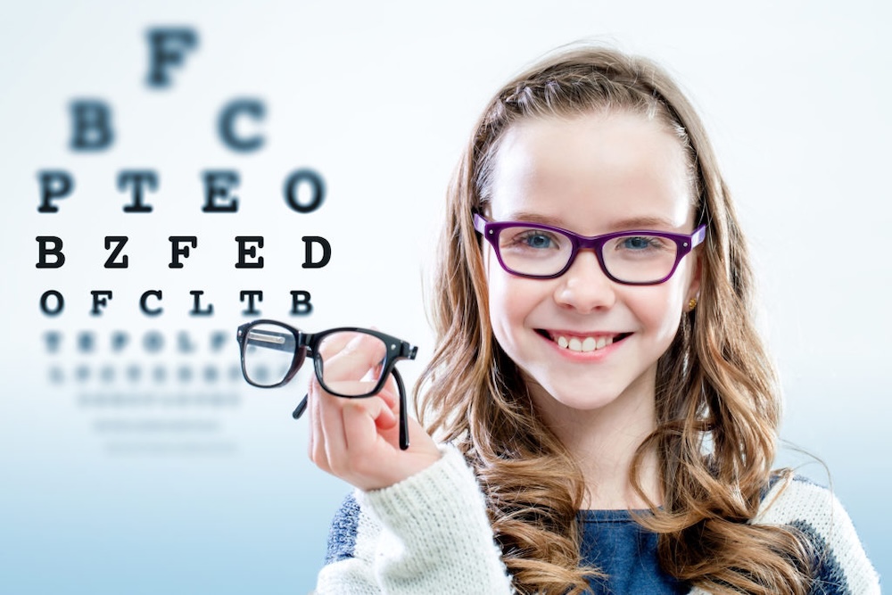 A little girl holding up a pair of glasses, while wearing glasses and smiling